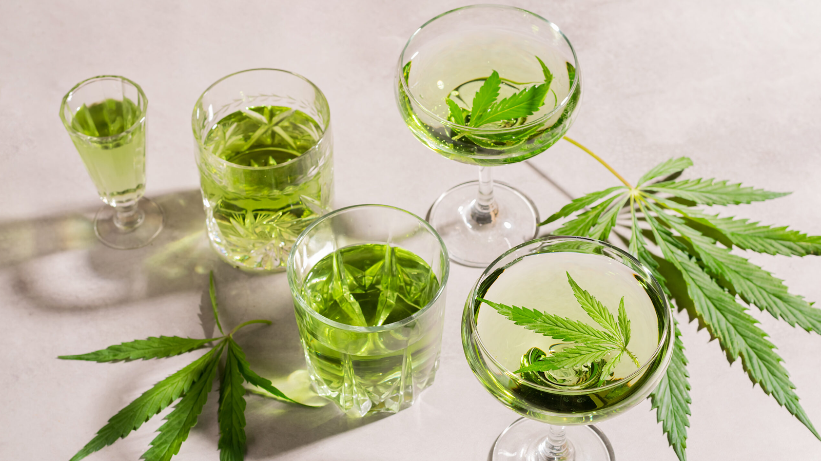 Various glasses of pale green cannabis-infused beverages garnished with fresh leaves on white surface