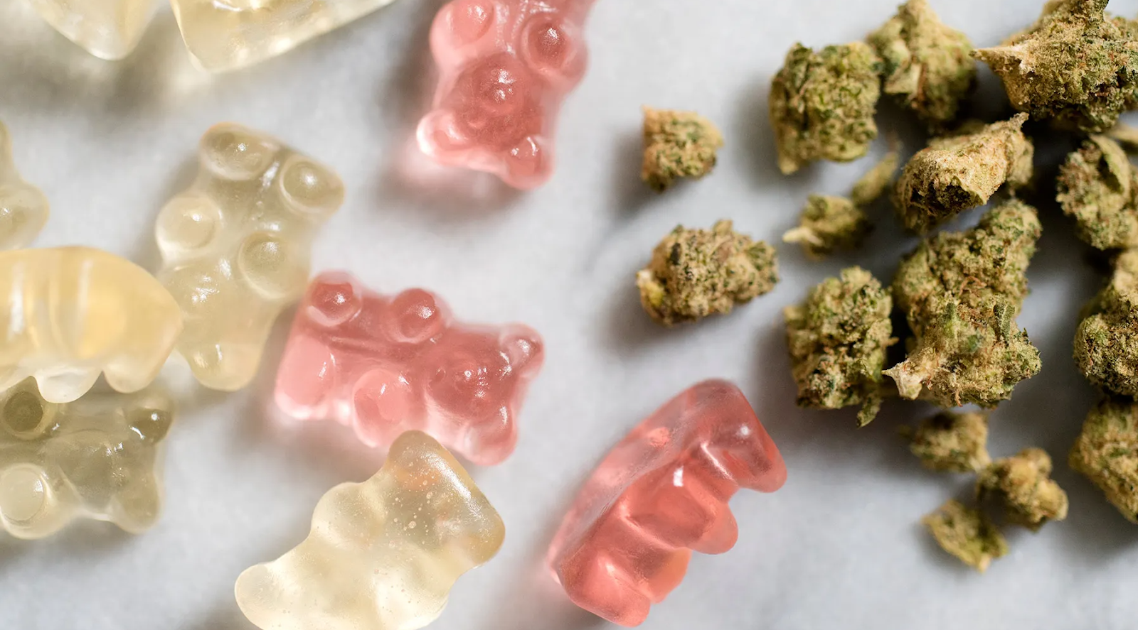 Clear and pink gummy bears next to cannabis flower buds on white surface
